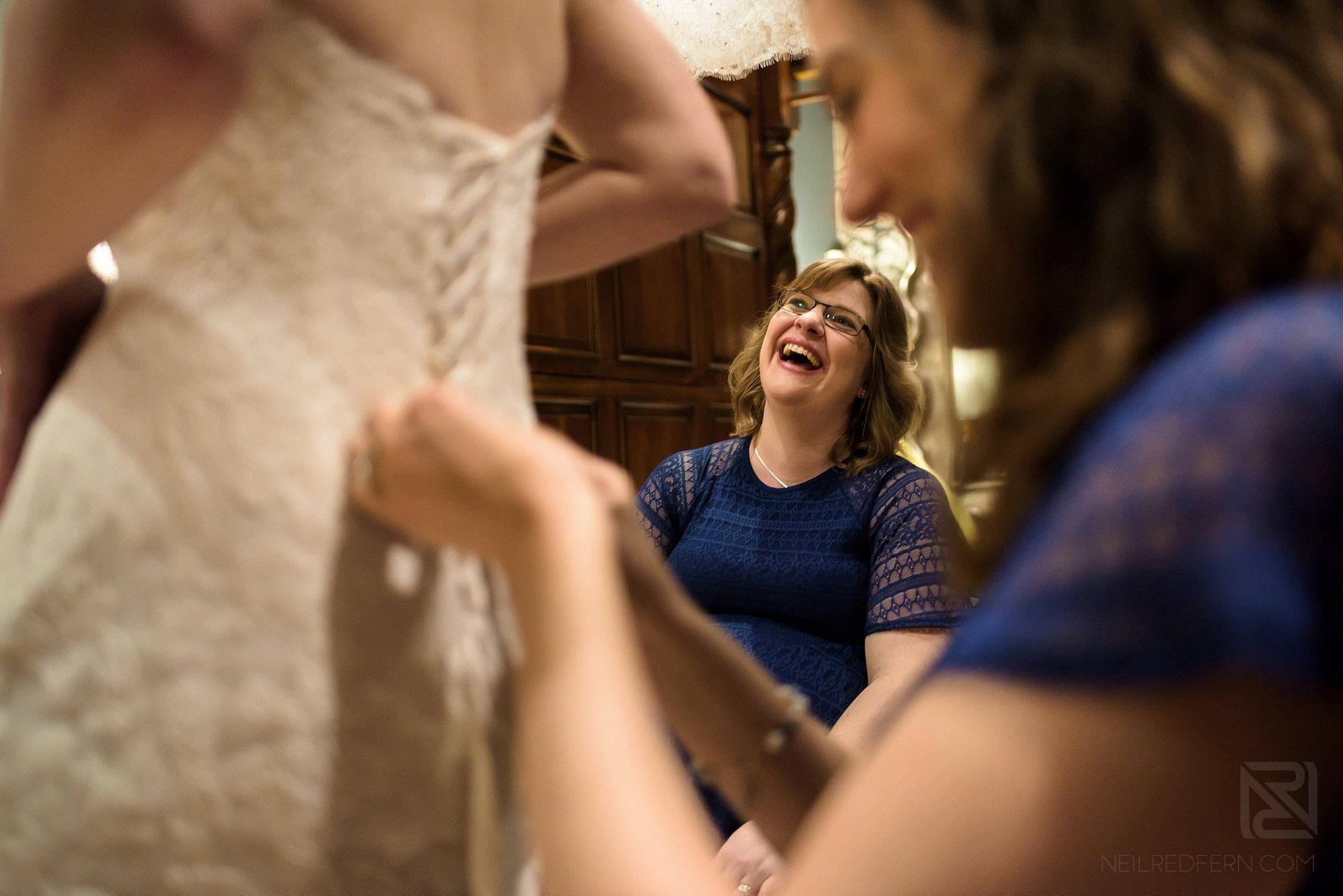 bridesmaids laughing putting bride in to wedding dress