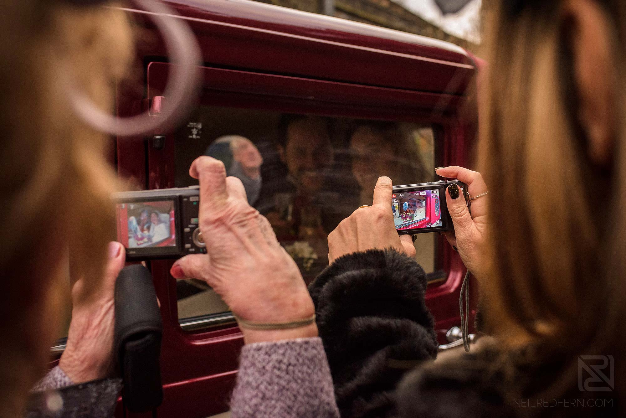 guests taking photograph of newlyweds in car