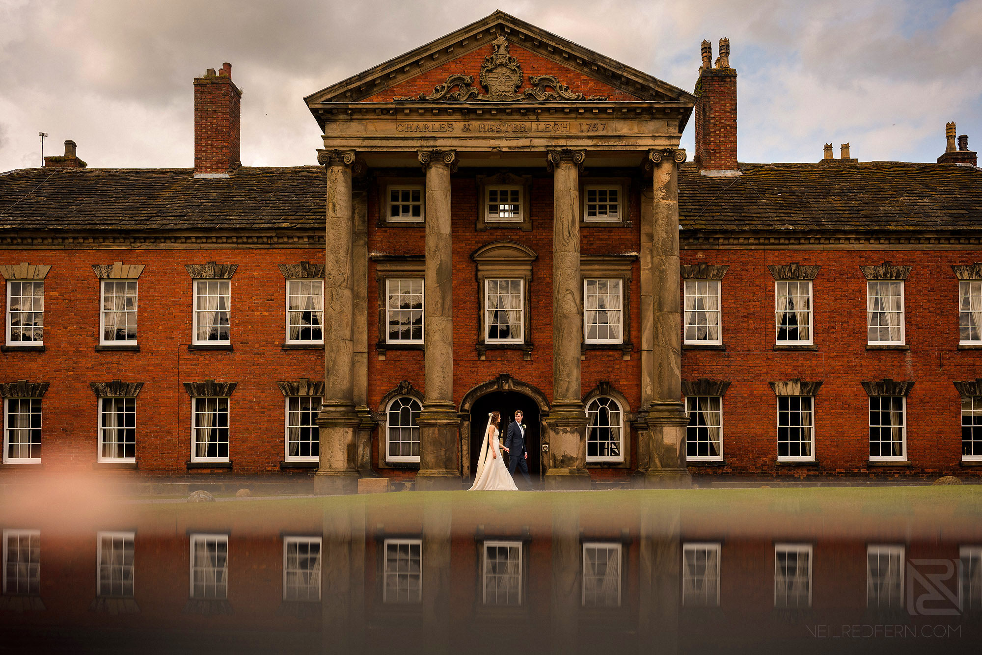 wide angle photograph of bride and groom walking past Adlington Hall in Cheshire